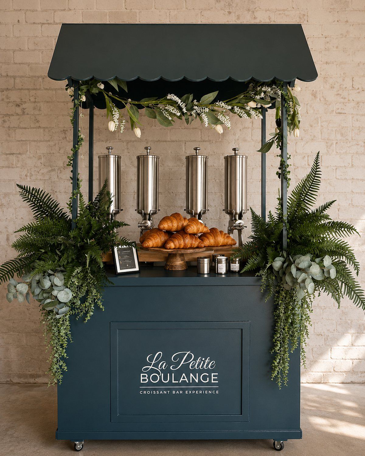 The La Petite Boulange signature croissant cart — a deep-green pastry cart with scalloped canopy, eucalyptus and fern styling, four stainless pastry injectors and a wooden stand of fresh croissants